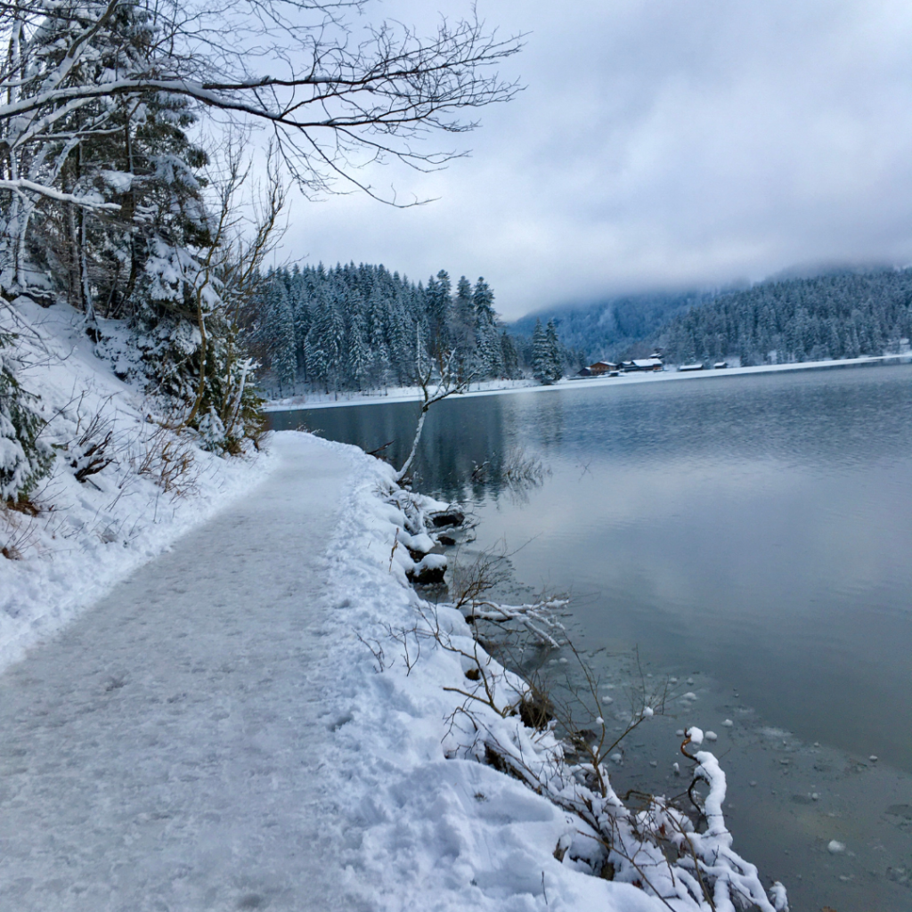 Verschneiter Fußweg direkt am Ufer eines Sees in Gebirgslandschaft.